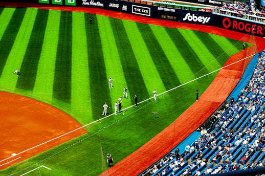 A baseball field with players in uniform stands ready near the sideline on the lush green grass. The stadium is partially filled with spectators in the seating area, and various advertisements are displayed on the perimeter fence.