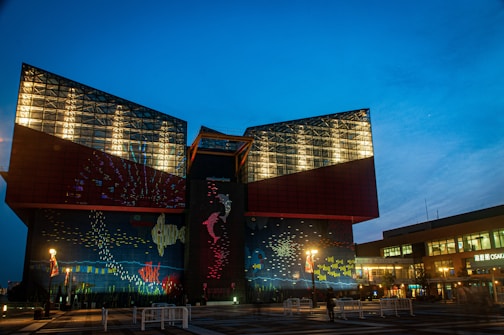 A large, modern aquarium building illuminated in the evening, featuring a colorful facade with depictions of marine life. The structure has a distinctive geometric design, with glass and red paneling. The surrounding area is lit with street lamps, and a few people are visible as silhouettes.