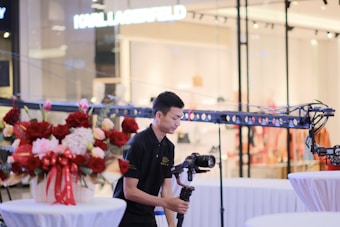 A person operating a camera stabilizer is positioned next to a table decorated with an arrangement of red and pink flowers tied with red ribbons. The setting seems to be indoors, likely in a shopping or event space with a Karl Lagerfeld sign visible in the background. Soft ambient lighting illuminates the scene with clean white tables in the foreground.