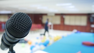 A close-up view of a microphone in the foreground with a blurred background of a room. The room contains colorful balloons scattered on the floor, suggesting an event or celebration setting. The background is out of focus, displaying some chairs and a person standing.
