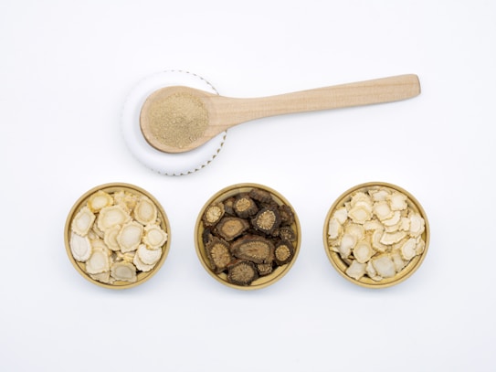 Three small round wooden bowls filled with various dried root slices are neatly arranged side by side. Above them, a wooden spoon rests on a ceramic dish, containing a small pile of beige powder. The items are placed on a clean white surface.