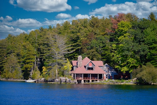 A quaint lakeside house is nestled among lush green trees under a vibrant blue sky with scattered clouds. The building features a stone chimney and a red-roofed design, creating a rustic and cozy appearance. The surrounding forest and clear water enhance the idyllic and serene atmosphere.