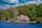 A quaint lakeside house is nestled among lush green trees under a vibrant blue sky with scattered clouds. The building features a stone chimney and a red-roofed design, creating a rustic and cozy appearance. The surrounding forest and clear water enhance the idyllic and serene atmosphere.