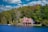 A quaint lakeside house is nestled among lush green trees under a vibrant blue sky with scattered clouds. The building features a stone chimney and a red-roofed design, creating a rustic and cozy appearance. The surrounding forest and clear water enhance the idyllic and serene atmosphere.