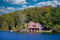 A quaint lakeside house is nestled among lush green trees under a vibrant blue sky with scattered clouds. The building features a stone chimney and a red-roofed design, creating a rustic and cozy appearance. The surrounding forest and clear water enhance the idyllic and serene atmosphere.
