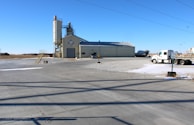 A concrete plant setup is visible with a large gray industrial building. A tall silo stands adjacent, featuring a logo. Several trucks, including a cement mixer and a freight truck, are parked nearby. Wide open spaces and a few scattered patches of snow surround the area under a clear blue sky.
