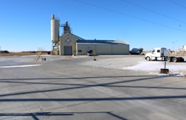 A concrete plant setup is visible with a large gray industrial building. A tall silo stands adjacent, featuring a logo. Several trucks, including a cement mixer and a freight truck, are parked nearby. Wide open spaces and a few scattered patches of snow surround the area under a clear blue sky.
