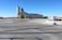 A concrete plant setup is visible with a large gray industrial building. A tall silo stands adjacent, featuring a logo. Several trucks, including a cement mixer and a freight truck, are parked nearby. Wide open spaces and a few scattered patches of snow surround the area under a clear blue sky.