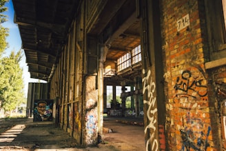 An abandoned industrial building with extensive graffiti on the walls. The structure shows signs of decay, with overgrown vegetation visible outside. Sunlight streams through large windows, casting shadows across the floor. A painted mural of a human face is visible in the distance.