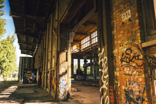 An abandoned industrial building with extensive graffiti on the walls. The structure shows signs of decay, with overgrown vegetation visible outside. Sunlight streams through large windows, casting shadows across the floor. A painted mural of a human face is visible in the distance.