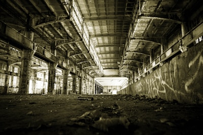 A large, empty industrial warehouse with high ceilings and rows of concrete support beams. The space is poorly lit, showing deteriorated walls with visible graffiti. The floor is covered with debris and dirt.