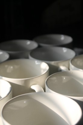 A collection of off-white ceramic mugs is arranged in close proximity. The lighting casts soft shadows and highlights on the mugs, emphasizing their smooth surfaces and round shapes.