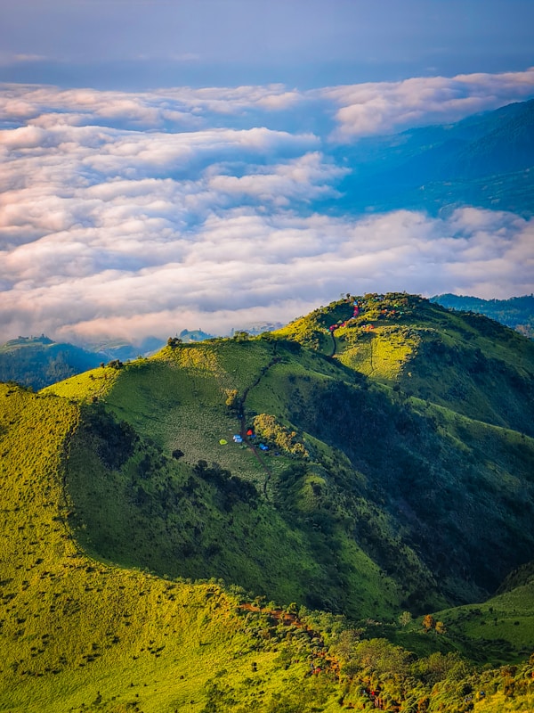 A lush, green mountain landscape with several colorful tents scattered amidst the greenery. The mountain range is covered with grass and sparse trees, giving way to an expansive cloud-filled sky. The clouds appear thick and dense, creating a serene and peaceful atmosphere.