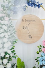 A wooden circular sign displays the words 'The Engagement of Ola & Ikei' surrounded by a floral arrangement. The flowers are a mix of white, pink, and blue blossoms set against green foliage. The blue flowers are prominently placed near the top right.