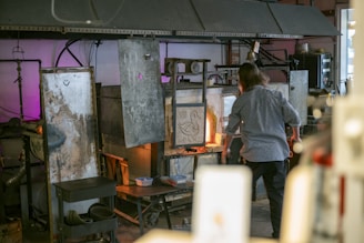 A person is working at a glassblowing studio. The setting includes several industrial pieces of equipment used for heating and shaping glass. The area is filled with various metal surfaces, some showing signs of wear and inscriptions. The space is dimly lit with a warm glow from the furnace, and there are tools and materials on a nearby cart.