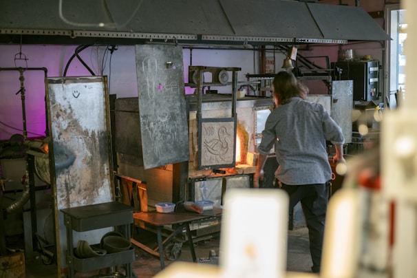 A person is working at a glassblowing studio. The setting includes several industrial pieces of equipment used for heating and shaping glass. The area is filled with various metal surfaces, some showing signs of wear and inscriptions. The space is dimly lit with a warm glow from the furnace, and there are tools and materials on a nearby cart.