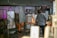 A person is working at a glassblowing studio. The setting includes several industrial pieces of equipment used for heating and shaping glass. The area is filled with various metal surfaces, some showing signs of wear and inscriptions. The space is dimly lit with a warm glow from the furnace, and there are tools and materials on a nearby cart.