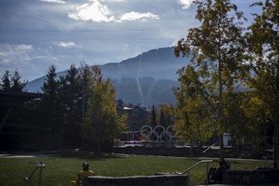 A scenic view captures a mountainous landscape under a partly cloudy sky. Shaded trees frame an open grassy area, leading to five interlocking rings, the iconic symbol of the Olympic Games. The mountain in the background is softly illuminated by diffuse sunlight.