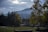 A scenic view captures a mountainous landscape under a partly cloudy sky. Shaded trees frame an open grassy area, leading to five interlocking rings, the iconic symbol of the Olympic Games. The mountain in the background is softly illuminated by diffuse sunlight.