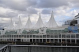 A large waterfront building with distinctive white sail-like structures on the roof, identified as Canada Place. The structure is adjacent to a body of water, with a cloudy sky in the background. The building's facade features glass windows and there is a mural visible on the side.