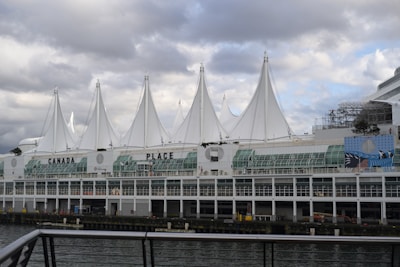 A large waterfront building with distinctive white sail-like structures on the roof, identified as Canada Place. The structure is adjacent to a body of water, with a cloudy sky in the background. The building's facade features glass windows and there is a mural visible on the side.