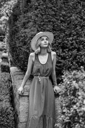 A young woman with shoulder-length hair is wearing a sleeveless long dress and a wide-brimmed straw hat. She is leaning against a stone wall with dense foliage behind her, suggesting a garden setting. The image is in black and white, emphasizing textures and lighting contrasts.