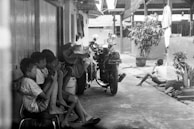 A group of children sit closely together on a narrow alleyway, possibly engaging with handheld objects. Nearby, a motorcycle is parked against the wall, surrounded by casually placed items and foliage in pots. The walls and roofs of small buildings create an enclosed atmosphere.