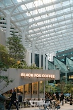 A modern coffee shop with a glass canopy overhead, surrounded by tall buildings in an urban setting. People sit and walk around the cafe, which is decorated with greenery and white furniture. The sign reads 'Black Fox Coffee' in illuminated letters.