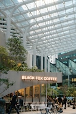 A modern coffee shop with a glass canopy overhead, surrounded by tall buildings in an urban setting. People sit and walk around the cafe, which is decorated with greenery and white furniture. The sign reads 'Black Fox Coffee' in illuminated letters.