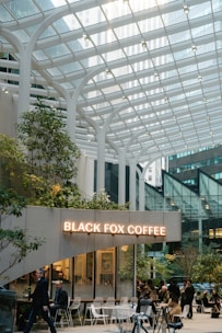 A modern coffee shop with a glass canopy overhead, surrounded by tall buildings in an urban setting. People sit and walk around the cafe, which is decorated with greenery and white furniture. The sign reads 'Black Fox Coffee' in illuminated letters.