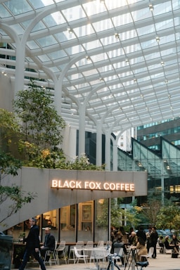 A modern coffee shop with a glass canopy overhead, surrounded by tall buildings in an urban setting. People sit and walk around the cafe, which is decorated with greenery and white furniture. The sign reads 'Black Fox Coffee' in illuminated letters.