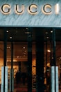 Glass storefront with sleek black doors, showcasing a luxurious and elegantly lit interior. The iconic brand name is prominently displayed above the entrance, set against a marble facade. Inside, there are upscale displays, plush furniture, and subtle lighting creating an opulent atmosphere.