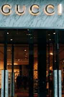 Glass storefront with sleek black doors, showcasing a luxurious and elegantly lit interior. The iconic brand name is prominently displayed above the entrance, set against a marble facade. Inside, there are upscale displays, plush furniture, and subtle lighting creating an opulent atmosphere.
