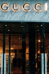 Glass storefront with sleek black doors, showcasing a luxurious and elegantly lit interior. The iconic brand name is prominently displayed above the entrance, set against a marble facade. Inside, there are upscale displays, plush furniture, and subtle lighting creating an opulent atmosphere.