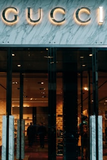 Glass storefront with sleek black doors, showcasing a luxurious and elegantly lit interior. The iconic brand name is prominently displayed above the entrance, set against a marble facade. Inside, there are upscale displays, plush furniture, and subtle lighting creating an opulent atmosphere.