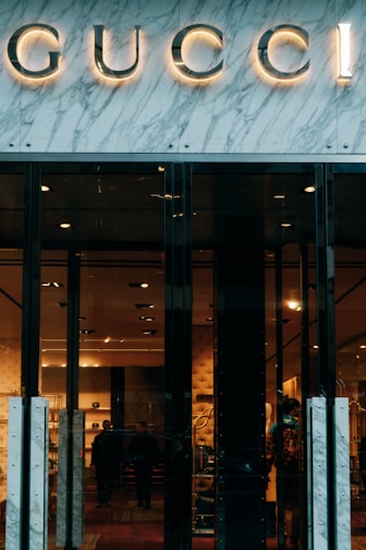 Glass storefront with sleek black doors, showcasing a luxurious and elegantly lit interior. The iconic brand name is prominently displayed above the entrance, set against a marble facade. Inside, there are upscale displays, plush furniture, and subtle lighting creating an opulent atmosphere.