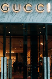 Glass storefront with sleek black doors, showcasing a luxurious and elegantly lit interior. The iconic brand name is prominently displayed above the entrance, set against a marble facade. Inside, there are upscale displays, plush furniture, and subtle lighting creating an opulent atmosphere.
