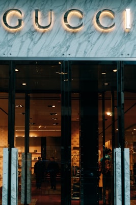 Glass storefront with sleek black doors, showcasing a luxurious and elegantly lit interior. The iconic brand name is prominently displayed above the entrance, set against a marble facade. Inside, there are upscale displays, plush furniture, and subtle lighting creating an opulent atmosphere.