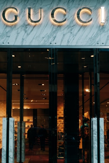 Glass storefront with sleek black doors, showcasing a luxurious and elegantly lit interior. The iconic brand name is prominently displayed above the entrance, set against a marble facade. Inside, there are upscale displays, plush furniture, and subtle lighting creating an opulent atmosphere.