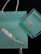 A Tiffany & Co. shopping bag and a matching box rest against a dark background. The box is open, revealing a silver necklace with a heart-shaped pendant. The iconic Tiffany blue color is prominent in both the bag and the box, complemented by white ribbon handles on the bag.