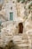 An ancient stone structure is built into a rocky hillside, featuring a unique stone façade with a weathered green wooden door. The staircase leading to the entrance is made of roughly hewn stone steps. Sparse vegetation grows around the entrance, adding a hint of greenery to the rustic setting.
