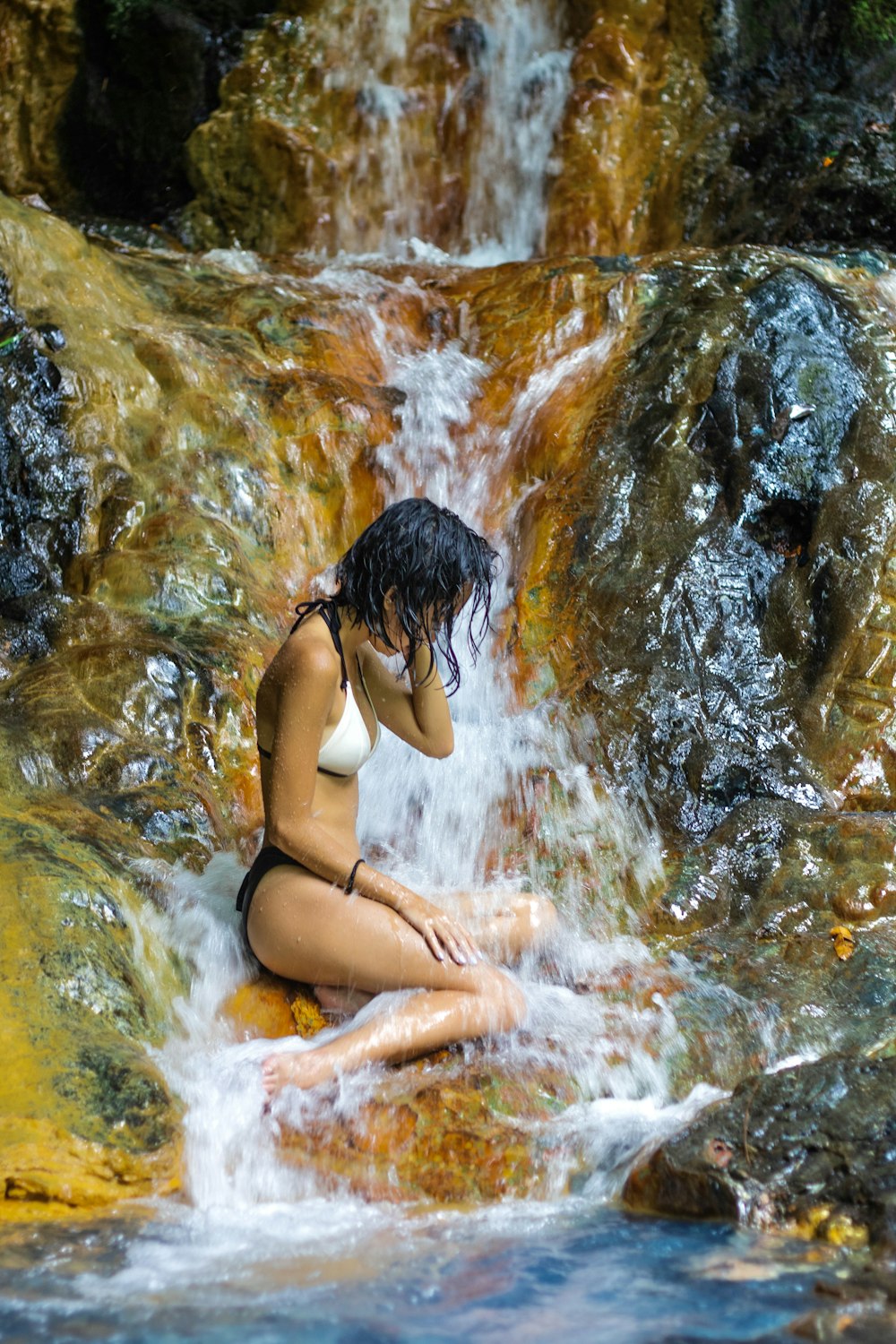 a woman in a bikini sitting on a rock next to a waterfall