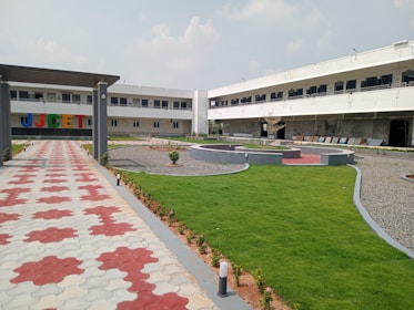 A two-story building with an L-shaped design is visible. The front has large colorful letters on the wall. There is a neatly manicured lawn area with sections of paving stones and gravel. The pathway leading to the building features a red and white pattern. The sky is partly cloudy, adding to the serene atmosphere.