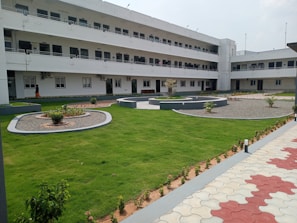A modern, multi-story building with a white facade and numerous windows is surrounded by a well-manicured lawn and landscaped flower beds. The central courtyard features pathways and neatly trimmed grass, while a person walks along the building.