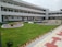 A modern, multi-story building with a white facade and numerous windows is surrounded by a well-manicured lawn and landscaped flower beds. The central courtyard features pathways and neatly trimmed grass, while a person walks along the building.