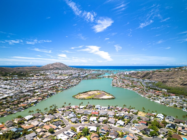 Aerial view of a coastal residential area with houses and roads lining a narrow green waterfront. A small island is situated in the waterway, surrounded by palm trees. In the background, mountains rise against a vibrant blue sky, with the ocean visible in the distance.