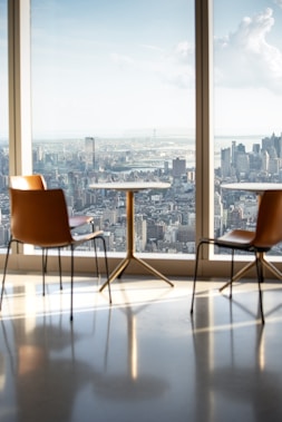A modern, minimalist interior with chairs and a table placed near large floor-to-ceiling windows overlooking a cityscape. The urban view outside is expansive, showcasing a variety of skyscrapers under a clear sky.