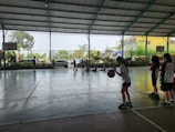 A group of young people are inside an indoor sports court. Several children are engaged in activity, with some playing basketball and others sitting on the floor against the fence. Greenery is visible outside through the netting.