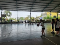 A group of young people are inside an indoor sports court. Several children are engaged in activity, with some playing basketball and others sitting on the floor against the fence. Greenery is visible outside through the netting.