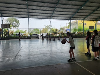 A group of young people are inside an indoor sports court. Several children are engaged in activity, with some playing basketball and others sitting on the floor against the fence. Greenery is visible outside through the netting.